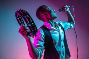 African American handsome jazz musician playing tambourine and singing into the microphone in the studio on a neon background. Music concept. Young joyful attractive guy improvising. Close-up retro portrait. Design By Freepik